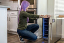 Person with purple hair kneeling next to a wine cooler in a kitchen.