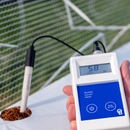A hand holding a BlueLab soil pH meter with a probe inserted into soil, with a blurred outdoor background and green netting behind it.