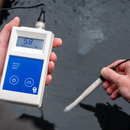 A hand holds a white and blue Biuetab soil pH meter with a probe connected, while another hand positions a separate pH probe against wet dark ground.